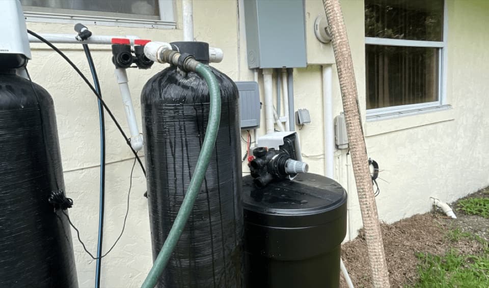 Black tank with a control valve and a green hose connected, mounted against a stucco wall near an electrical panel and a window
