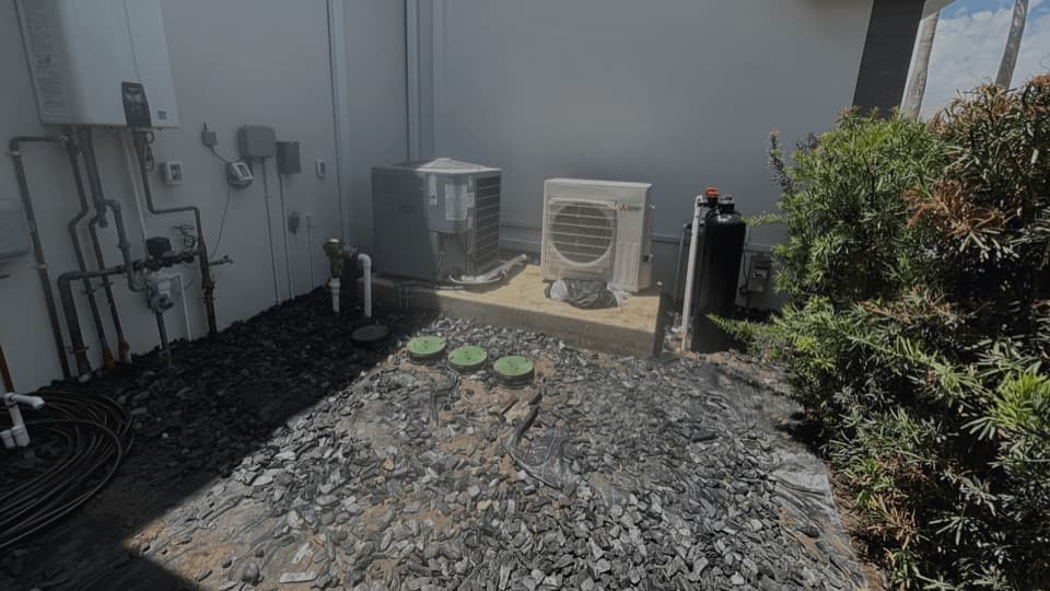 Condenser units, a black tank, coiled black tubing and three green utility caps set in gravel against a building wall