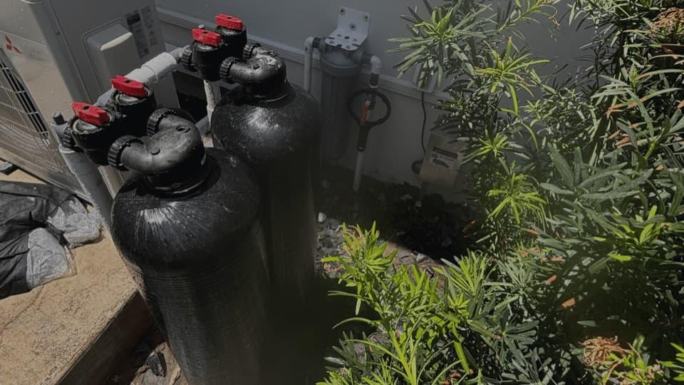 Two black tanks with red valve handles against a building wall with a blue filter housing, surrounded by shrubs