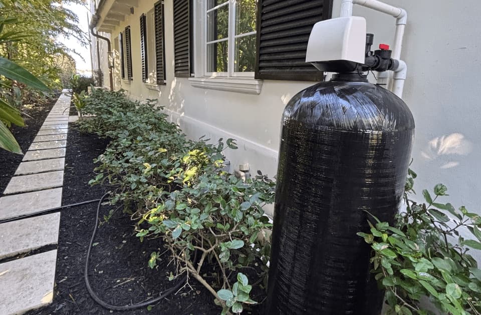 Black tank with a white valve head against a home exterior with dark shutters, next to a stone walkway and shrubs