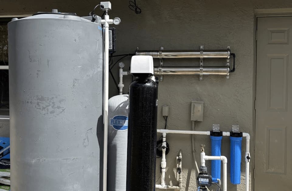 A large grey tank, a white tank, a black tank with a white valve head and horizontal filter cartridges mounted on a stucco wall near a door