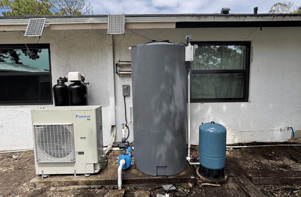 Grey water storage tank with a solar panel and pressure gauge, twin black tanks, a condenser unit and a blue pressure tank against a house exterior